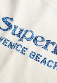 Niebieski tekst wysadzany kryształkami w kształcie słowa "Superior" i "Venice Beach" na białym materiale.