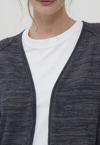 Tummanharmaa neulottu neuletakki, jossa on sileä pinta ja avonainen etuosa, käytetty valkoisen pyöreäkauluksisen t-paidan päällä. Hillitty kuvioteema koko vaatteessa.