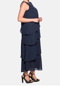 Navy blue tiered maxi dress with layered chiffon fabric, beaded neckline, and high-heeled black sandals with ankle strap.