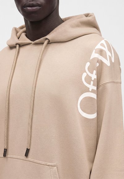 Personne à la peau foncée portant un sweat à capuche beige avec un texte partiel blanc sur l'épaule droite et des cordons qui pendent.