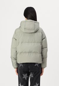 Doudoune grise avec capuche, comportant des sections matelassées et un design cropped. Les coutures visibles renforcent la texture. Portée avec un pantalon à motifs étoiles.