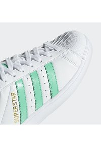 Baskets en cuir blanc avec trois rayures vertes, bout renforcé texturé et lettrage doré "SUPERSTAR" sur le côté. Semelle en caoutchouc et design à lacets.