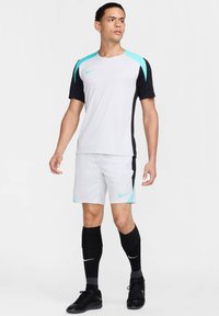 Nike sportkleding set bestaande uit een wit bovenstuk met zwarte mouwen en turquoise accenten, gecombineerd met witte shorts en zwarte kniekousen.