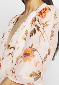 Blouse légère et transparente avec un décolleté en V profond, ornée d'un motif floral orange et jaune sur un fond pâle. Manches courtes et texture douce.
