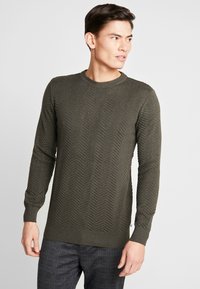 Olivgrüner Strickpullover mit Zickzackmuster, Rundhalsausschnitt und gerippten Bündchen und Saum. Über karierten Hosen getragen.
