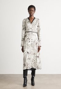 Long, patterned dress in cream with brown illustrations, featuring a V-neckline and a black belt. Worn with fitted black boots.