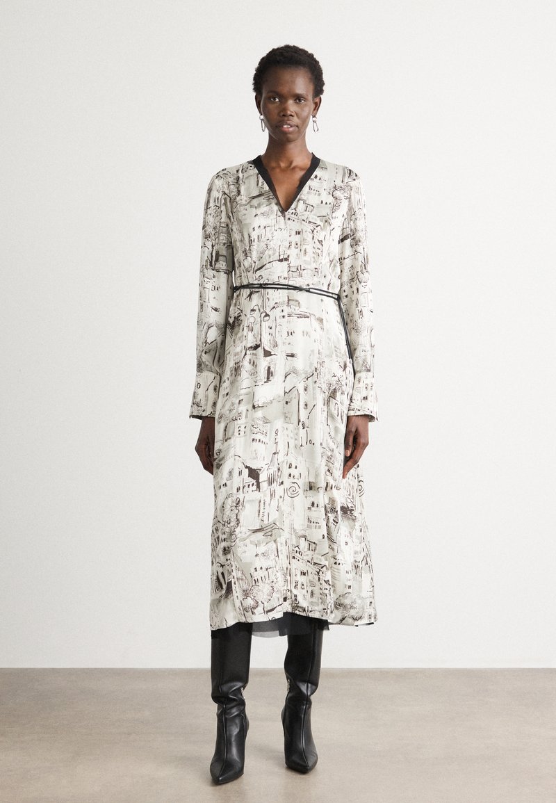 Long, patterned dress in cream with brown illustrations, featuring a V-neckline and a black belt. Worn with fitted black boots.