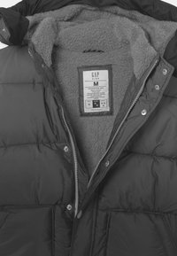 Chaqueta acolchada negra con forro de forro polar gris, cierres a presión, cremallera frontal y etiqueta visible que indica la talla M de GAP Kids.