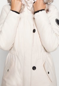 Cream-colored winter coat featuring a fur-lined hood, black buttons, elastic waist, and side pockets with flaps. Smooth texture and warm insulation.