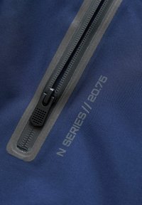 Marineblauwe stof met een gladde textuur, voorzien van een weerbestendige rits in donkergrijs en bedrukte "N SERIES // 20.76" details.