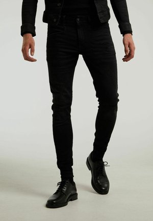 Mężczyzna ubrany w czarne, podarte obcisłe jeansy i czarne skórzane eleganckie buty, chodzący po jednolitym jasnym tle.