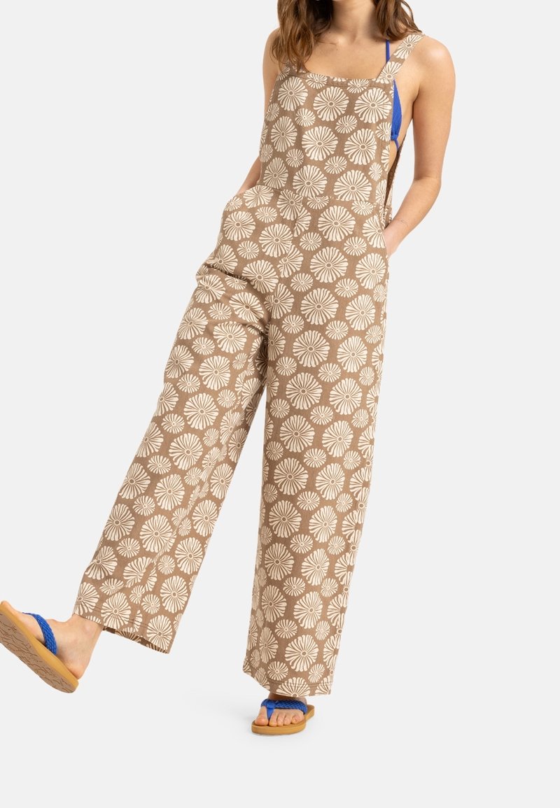 Combinaison en brown à motifs avec un design floral blanc, jambes larges et poches. Associée à des tongs bleues, mettant en valeur des lanières et une semelle texturée.
