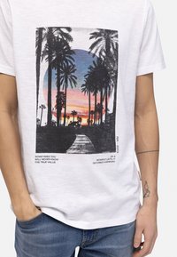 T-shirt en coton blanc avec un graphique de palmier en noir et en couleur, accompagné d'un coucher de soleil, avec du texte imprimé dans une police standard.