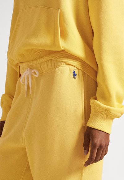 Rumena jopica s kapuco in sprednjim žepom ter rebrastimi manšetami, kombinirana z rumenimi trenirkami z elastičnim pasom in majhnim modrim logotipom.