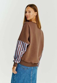 Hnedá mikina s rukávmi pruhovanými v modrej a bordovej farbe. Obsahuje kvetinové vzory na rukávoch a má voľný strih.