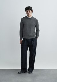 Moški črno-beli pulover z vzorcem, dolgimi rokavi in okroglim izrezom, kombiniran s prilagojenimi črnimi hlačami in črnimi čevlji na enobarvnem ozadju.