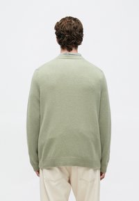 Homem com cabelo cacheado curto, vestindo um suéter de malha verde claro e calças bege, de costas contra um fundo branco simples.