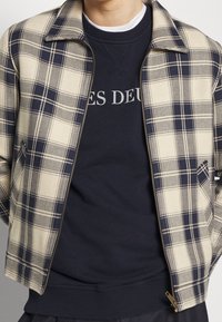 Chaqueta a cuadros beige y azul marino con cremallera frontal, cuello y dos bolsillos laterales, superpuesta sobre una sudadera azul marino con letras blancas.