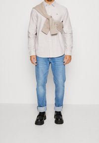 Lichtbeige overhemd met knopen, opgerolde mouwen, marineblauw logo. Lichtblauwe spijkerbroek, omgeslagen pijpen bij de enkels. Zwarte schoenen met dikke zolen. Trui los over de schouders gehangen.