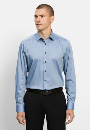 Mann trägt ein hellblaues Hemd mit Knöpfen, das in schwarze Hose mit schwarzem Gürtel gesteckt ist, schaut leicht nach links.