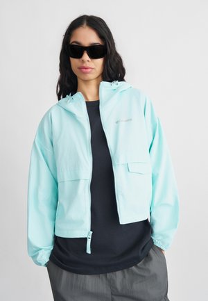 Žena nosící černé sluneční brýle, světle modrou bundu Columbia na zip, černé tričko a šedé kalhoty, stojící před jednobarevným bílým pozadím.