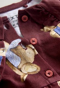 Chemise pour enfants bordeaux avec un imprimé d'ours, dotée d'un col et de boutons rouges. Le tissu a une texture douce avec des volants gonflés près du col.