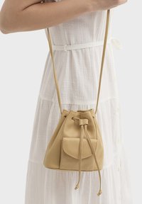 Sac seau en cuir beige avec fermeture à cordon et poche extérieure. Texture douce ; sangle courte et fine. Porté sur une robe blanche.
