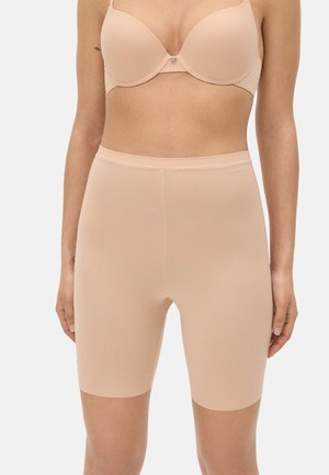 Mujer con shorts moldeadores nude de cintura alta sin costuras y sujetador nude a juego, de pie con los brazos relajados a los lados.