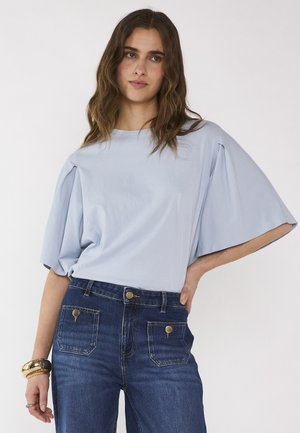 Femme aux cheveux bruns ondulés porte un chemisier bleu clair à manches larges, rentré dans un jean taille haute bleu avec des boutons dorés, accessoirisé avec des bracelets.