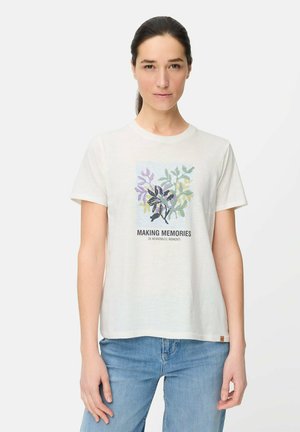 Kobieta o ciemnych włosach związanych w kucyk, ubrana w białą koszulkę z kwiatowym wzorem i napisem „TWORZENIE WSPOMNIEŃ W ZNACZĄCYCH CHWILACH”, w połączeniu z niebieskimi dżinsami.