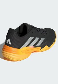 Černá atletická bota s prodyšným síťovaným svrškem, stříbrnými pruhy a oranžovou gumovou podrážkou. Má texturovaný vzor a pevné šněrování.