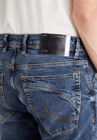 Jeans de denim azul-escuro com uma textura gasta, realçados por costuras laranja, apresentando um retalho de couro preto com logótipo e etiqueta na cintura.