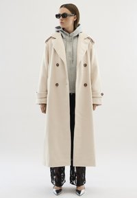Palton bej din lână cu un design cu două rânduri de nasturi și nasturi maro, purtat peste o hanorac gri cu fermoar și pantaloni negri din dantelă, completat cu pantofi ascuțiți argintii.