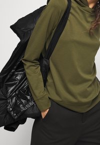 Osoba oblečená v olivově zeleném dlouhém rukávu a černých kalhotách, s jednou rukou v kapse, držící černou péřovou bundu přes rameno.