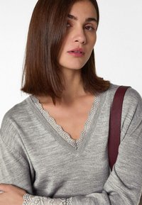 Grau V-Ausschnitt Pullover mit Spitze am Ausschnitt und den Bündchen. Weicher Stoff und lockere Passform, getragen mit einer bordeauxfarbenen Schultertasche.