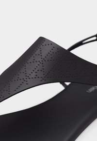 Κοντινό πλάνο του λουριού μαύρου σανδαλιού με διάτρητο μοτίβο αστεριού και σταυρού σε λείο υλικό.