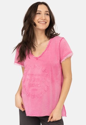 Young woman with long brown hair wearing a pink V-neck t-shirt with "Positive Life Positive Mind Club" text and hearts, smiling, hands in pockets.