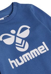 Blauwe katoenen langemouwen top met een witte logo van een bij-ontwerp en de merknaam "hummel" in vette letters. Ronde hals met drukknoopsluitingen.