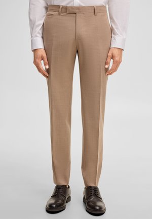 Mężczyzna ubrany w beżowe spodnie na miarę, białą koszulę i ciemnobrązowe skórzane eleganckie buty, stojący na jednolitym tle.