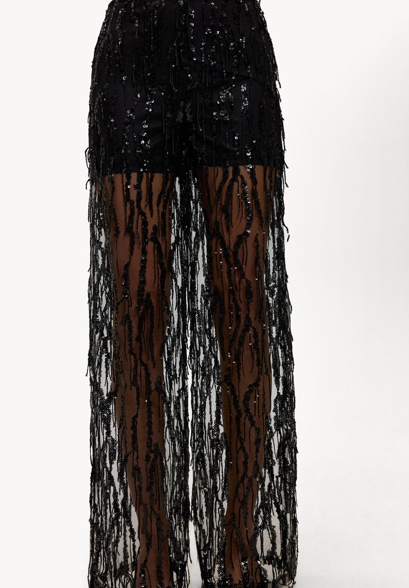 Pantalon noir à sequins avec un voile transparent orné de franges verticales. Le tissu est léger et semi-transparent, mettant en valeur l'aspect superposé.
