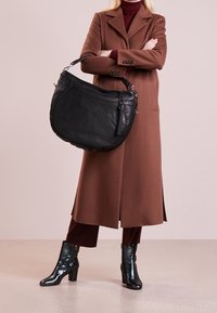 Czarna skórzana torebka z plecionymi akcentami, paskiem na ramię i detalem z frędzlami. Gładka tekstura i zakrzywiony kształt, noszona przez osobę w brązowym płaszczu.