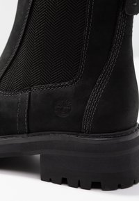 Musta mokkanahkainen nilkkaboots, jossa on elastiset sivupaneelit, halkiopinta, tikatut yksityiskohdat ja kumipohja, jossa on pito.