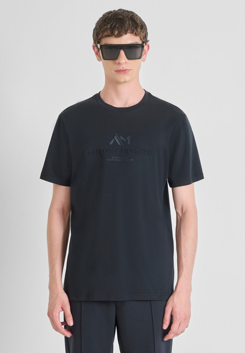 Camiseta de algodón negra con logo en relieve en el pecho, cuello redondo y mangas cortas, llevada con gafas de sol rectangulares.