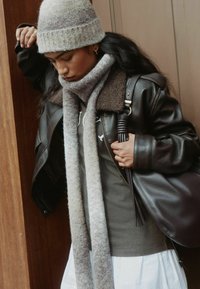 Brown leather jacket with shearling collar, grey knitted beanie, and matching scarf. White shirt, dark bag with textured finish.