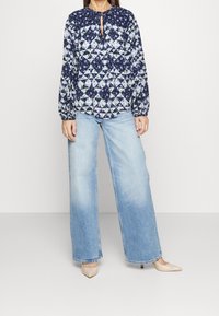 Langarmblouse met een marineblauw bloemen patchwork ontwerp, voorzien van een ronde halslijn en een sleufdetail. Gepaard met lichtblauwe wijde jeans.