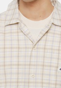 Uomo che indossa una camicia button-up a scacchi beige sopra una t-shirt bianca a girocollo, con il collo e la parte superiore del petto in vista.