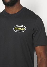 T-shirt noir en coton avec un logo oval "NIKE" en jaune et des accents en damier sur la poitrine gauche. Manches courtes, design à col rond.