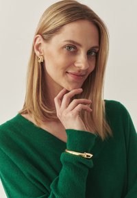Grüner Strickpullover mit V-Ausschnitt, getragen mit goldenen Ohrringen in abstrakter Form und einem goldenen Armband mit einer spitzen Akzentuierung.