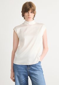 Hihaton, kerma väri blouse korkealla kaula-aukolla, valmistettu sileästä ja kevyestä kankaasta. Rentoa istuvuutta, jossa on hienovaraisia laskoksia.