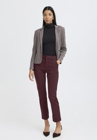 Blazer in houndstoothpatroon met een getailleerde pasvorm, gecombineerd met aangesloten bordeauxrode broek. Beide kledingstukken zijn gemaakt van geweven stof. Bruine puntige schoenen.